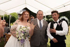 Bride and groom showing wedding rings with officiant under white tent, bride holding purple and white bouquet.