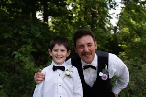 Smiling boy and groomsman in matching black bow ties and boutonnieres posing together at outdoor wedding.