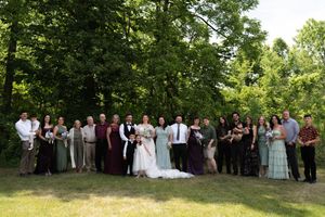 Large wedding party group photo in formal attire standing in front of lush green trees outdoors.