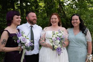 Wedding party smiling outdoors with bride holding purple and white bouquet flanked by bridesmaids and groom in green tie.