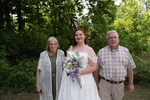 Bride in white off-shoulder gown holding purple bouquet poses with elderly couple outdoors near green trees.