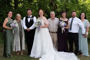 Bride and groom with family members in formal wedding attire posing outdoors among green trees.