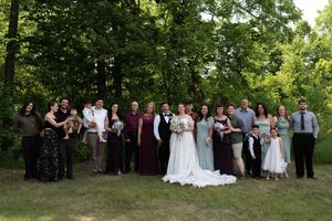 Large wedding party group photo on green lawn surrounded by lush trees in outdoor summer ceremony setting.