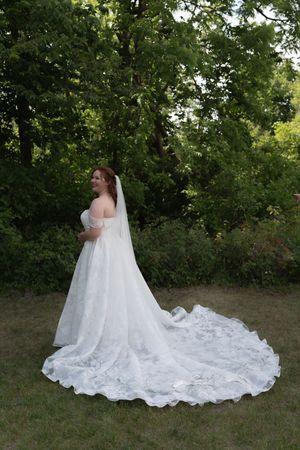 Bride in elegant lace wedding gown with long train and veil poses in a lush green garden setting.