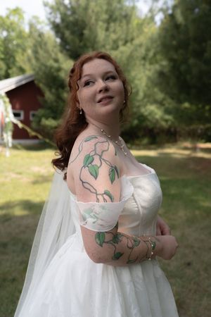 Tattooed bride in white off-shoulder gown with veil poses outdoors near red barn surrounded by green trees.