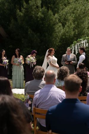 A bride in a white gown stands at an outdoor wedding altar with bridesmaids holding purple bouquets behind her.