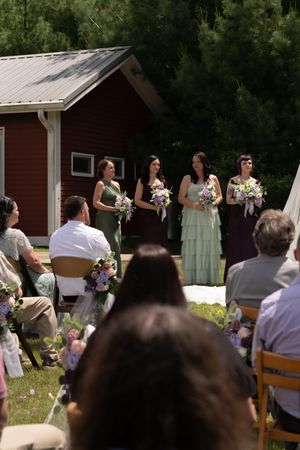 Four bridesmaids in sage green and purple gowns hold bouquets at an outdoor wedding ceremony near a red barn.