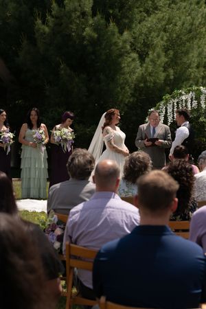 Outdoor wedding ceremony with bride in white gown and veil exchanging vows under floral arch with guests seated.