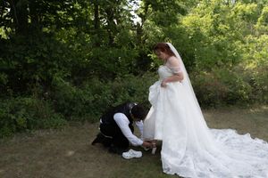 Groom kneeling to adjust brides wedding dress train in wooded outdoor setting before ceremony.