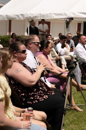 Wedding guests seated outdoors in sunny weather watching ceremony near a white tent on a summer day.