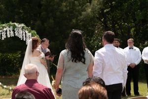 Outdoor wedding ceremony with bride in white veil and guests seated on sunny lawn near floral arch.