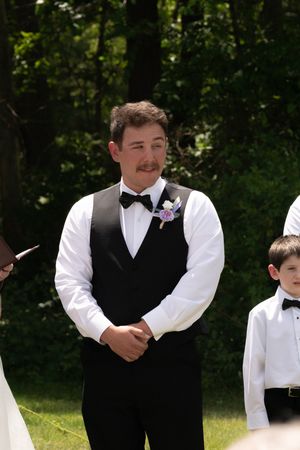 Groom in black vest and bow tie with boutonniere smiling during outdoor wedding ceremony.