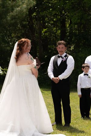 Bride in white gown reads vows to groom in black vest during an intimate outdoor wedding ceremony with greenery backdrop.