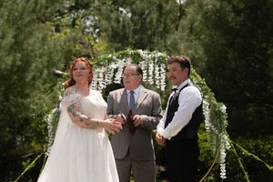 Bride and groom stand with officiant beneath a floral arch during an outdoor wedding ceremony surrounded by lush greenery.