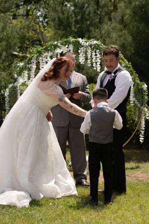 Bride and groom exchange vows with young ring bearer under floral arch at outdoor wedding ceremony.