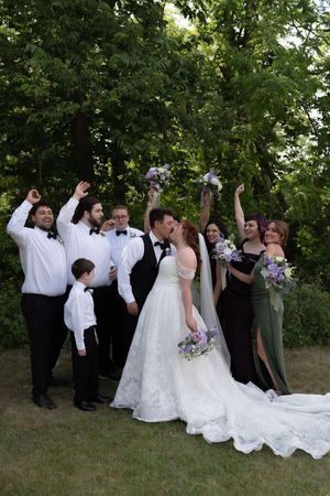 Wedding party celebrating with bride and groom kissing outdoors surrounded by lush green trees.
