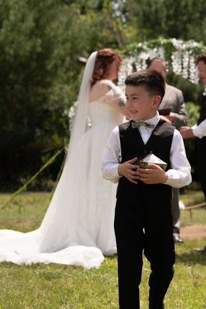 Smiling young boy in black vest and bow tie holds a gift box at an outdoor wedding ceremony.