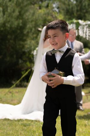 Happy young ring bearer in formal black vest and bow tie holds a small gift at an outdoor wedding.