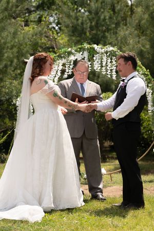 Bride and groom exchange vows outdoors under floral arch with officiant holding book on sunny day.