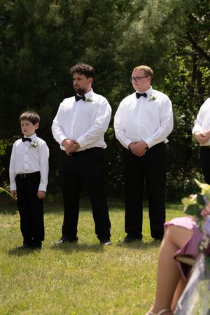 Three groomsmen in white shirts and black bow ties stand attentively during an outdoor wedding ceremony on green grass.