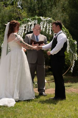 Bride and groom hold hands during outdoor wedding ceremony officiated under white floral circular arch.