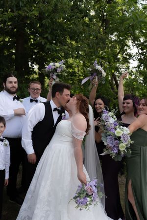 Bride and groom share a kiss surrounded by cheering wedding party holding purple bouquets in an outdoor setting.