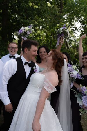 Joyful bride and groom almost kissing while wedding party celebrates behind them with bouquets raised in the air.