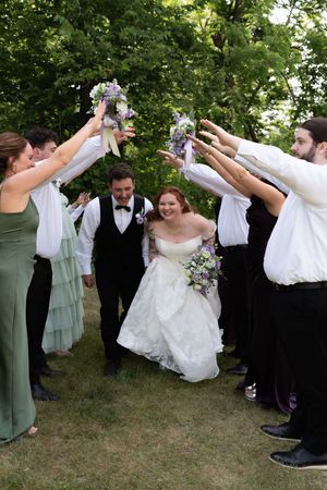 A bride and groom walk through an arch of raised hands formed by their wedding party in a lush green outdoor setting.