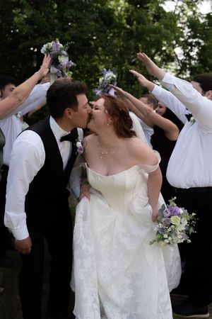 Bride and groom share first kiss under arch of raised bouquets held by wedding party outdoors.