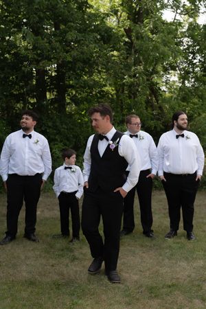Groom in black vest and bow tie stands with groomsmen in white shirts on green lawn near trees.