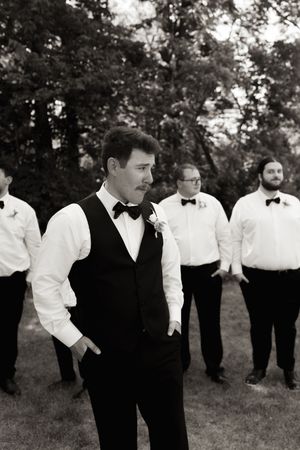 Black and white photo of groom in vest and bow tie standing before groomsmen outdoors.