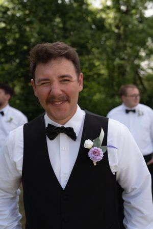 Smiling groom in black vest and bow tie with purple boutonniere at outdoor wedding ceremony.