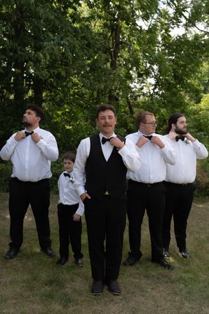 Groom and groomsmen adjusting bow ties outdoors in a fun posed wedding party photo with trees behind.