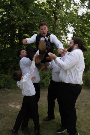Groomsmen in white shirts playfully lift the groom in a black vest into the air outdoors at a wedding.