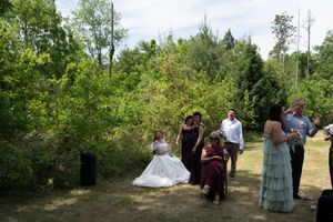 Wedding party gathering outdoors on grass with trees, including bride in white gown and guests in formal attire.
