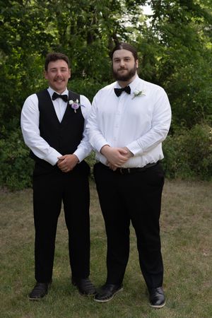 Two groomsmen in black vests and bow ties with boutonnieres posing outdoors at a wedding.
