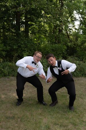 Two groomsmen in black bow ties striking fun poses on grass in front of trees at wedding.