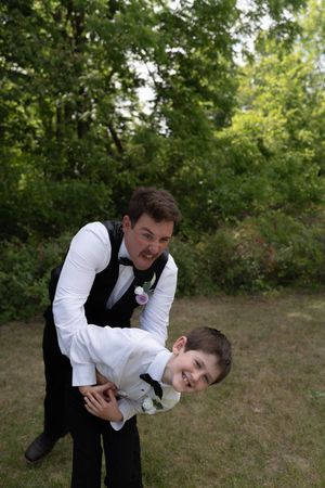 A groom in a black vest and bow tie playfully lifts a young boy in a white shirt outdoors at a wedding.