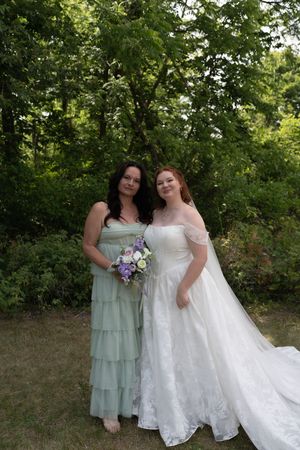 Bride in white lace gown stands with bridesmaid in sage green dress holding purple bouquet outdoors.