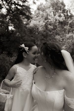 Black and white photo of bride in veil tenderly kissing flower girl on the cheek at outdoor wedding.