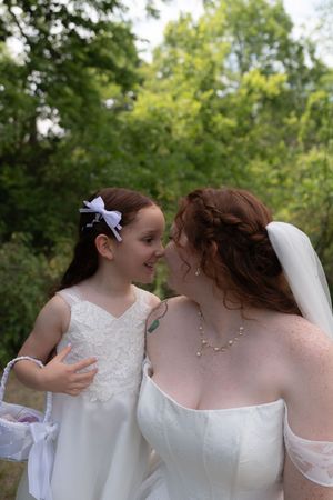 Bride in white gown shares a tender moment with flower girl in matching white dress at outdoor wedding.