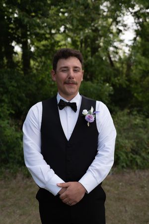 Handsome groomsman in black vest and bow tie with purple boutonniere posing outdoors at wedding.