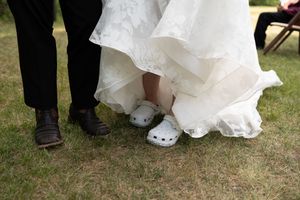 Close-up of bride wearing white Crocs under her wedding gown beside groom in brown dress shoes on grass.