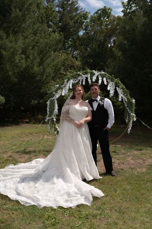 Bride and groom pose together under white wisteria floral arch on green lawn surrounded by trees.
