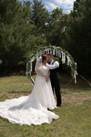 Bride and groom sharing a kiss under a floral arch adorned with white wisteria in a lush green outdoor setting.