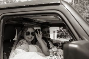 Black and white photo of smiling bride and groom wearing sunglasses inside a vehicle after wedding.