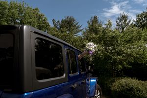 A hand holding a bridal bouquet of purple and white flowers extends from a blue Jeep surrounded by green trees.