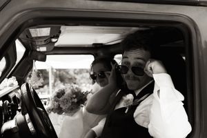 Black and white photo of bride and groom wearing sunglasses inside a vintage vehicle on wedding day.
