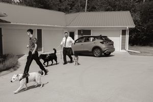 Black and white photo of two men walking dogs outside a building near a parked SUV.