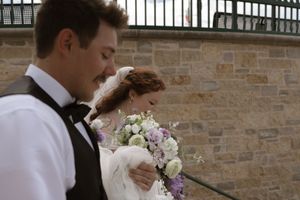 A bride holding a purple and white bouquet stands beside her groom near a stone wall.